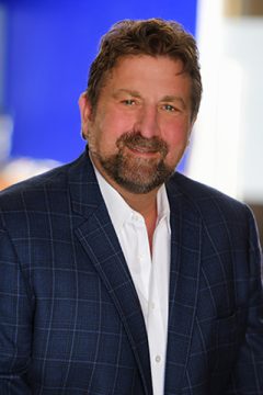 A bearded man named Leland William Gerber wearing a blue shirt, smiling and looking confidently at the camera.