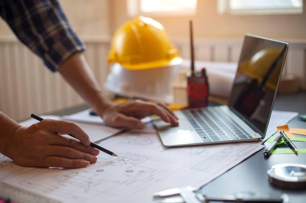 A man works on a laptop and draws on blueprints, highlighting a dynamic work environment