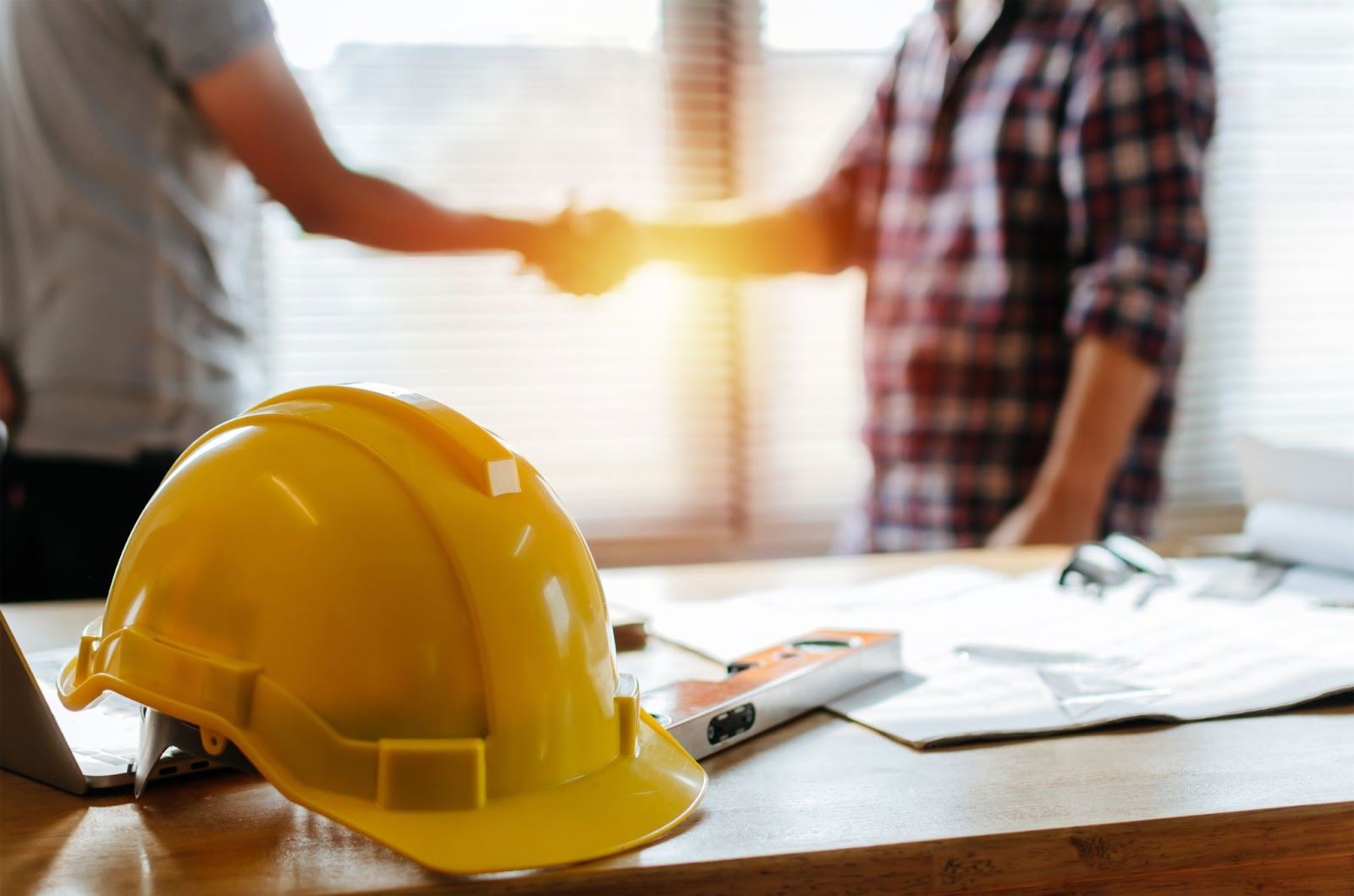 Two men in hard hats shake hands at a desk, symbolizing collaboration in commercial construction and expertise.