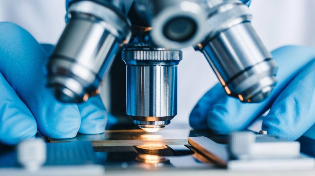 A scientist in blue gloves analyzes a specimen through a microscope