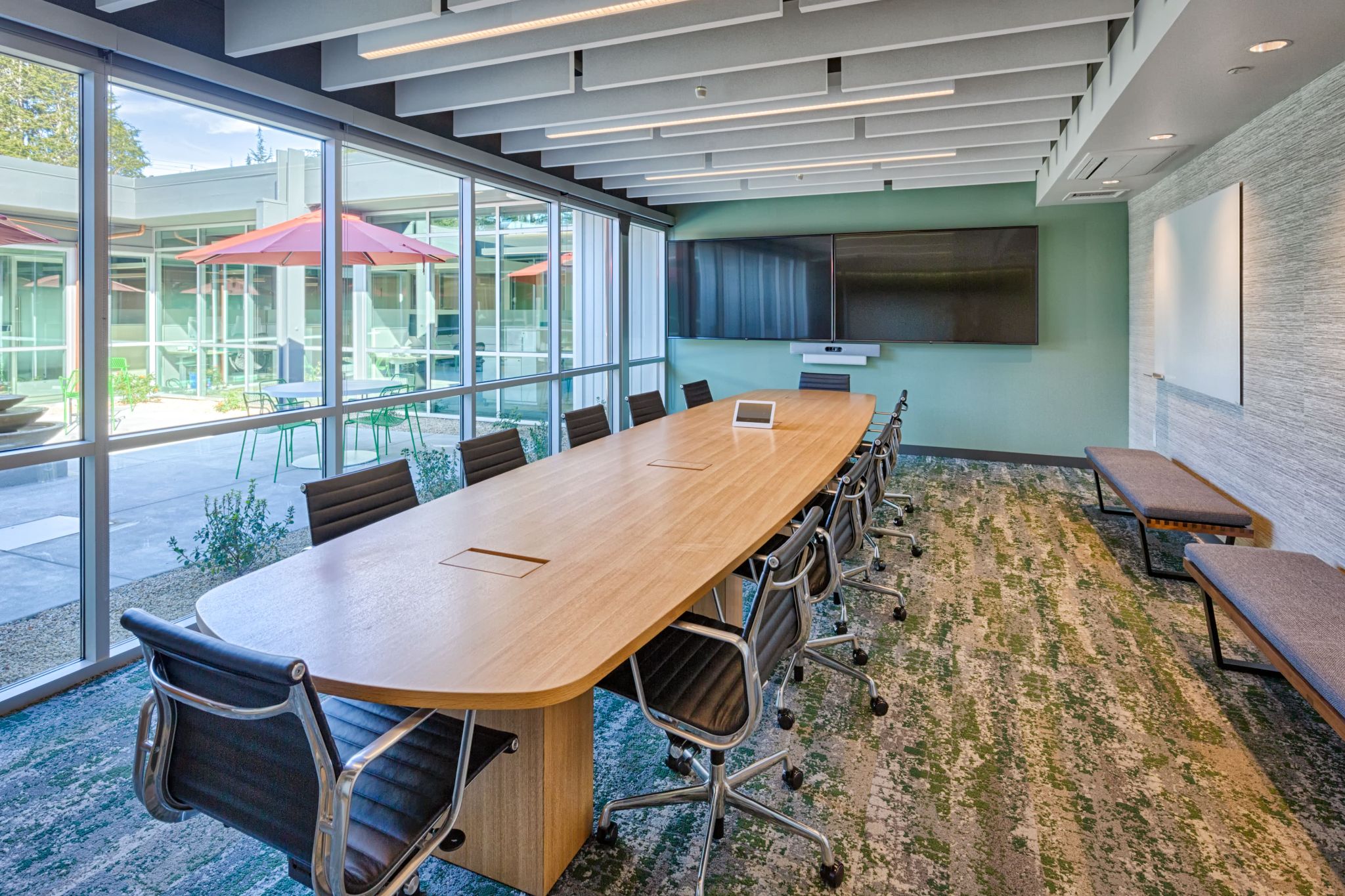 A modern conference room featuring a long wooden table, black office chairs, wall-mounted display screens, and floor-to-ceiling windows overlooking an outdoor courtyard with patio seating.