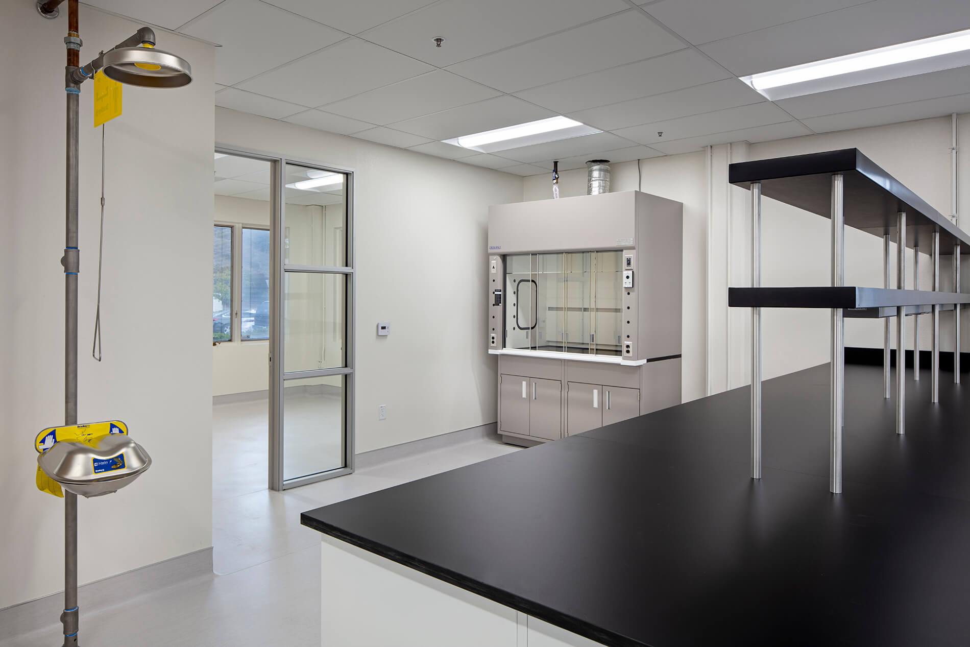An empty, modern laboratory with black countertops, shelving, a chemical fume hood, and an emergency eye washshower station.