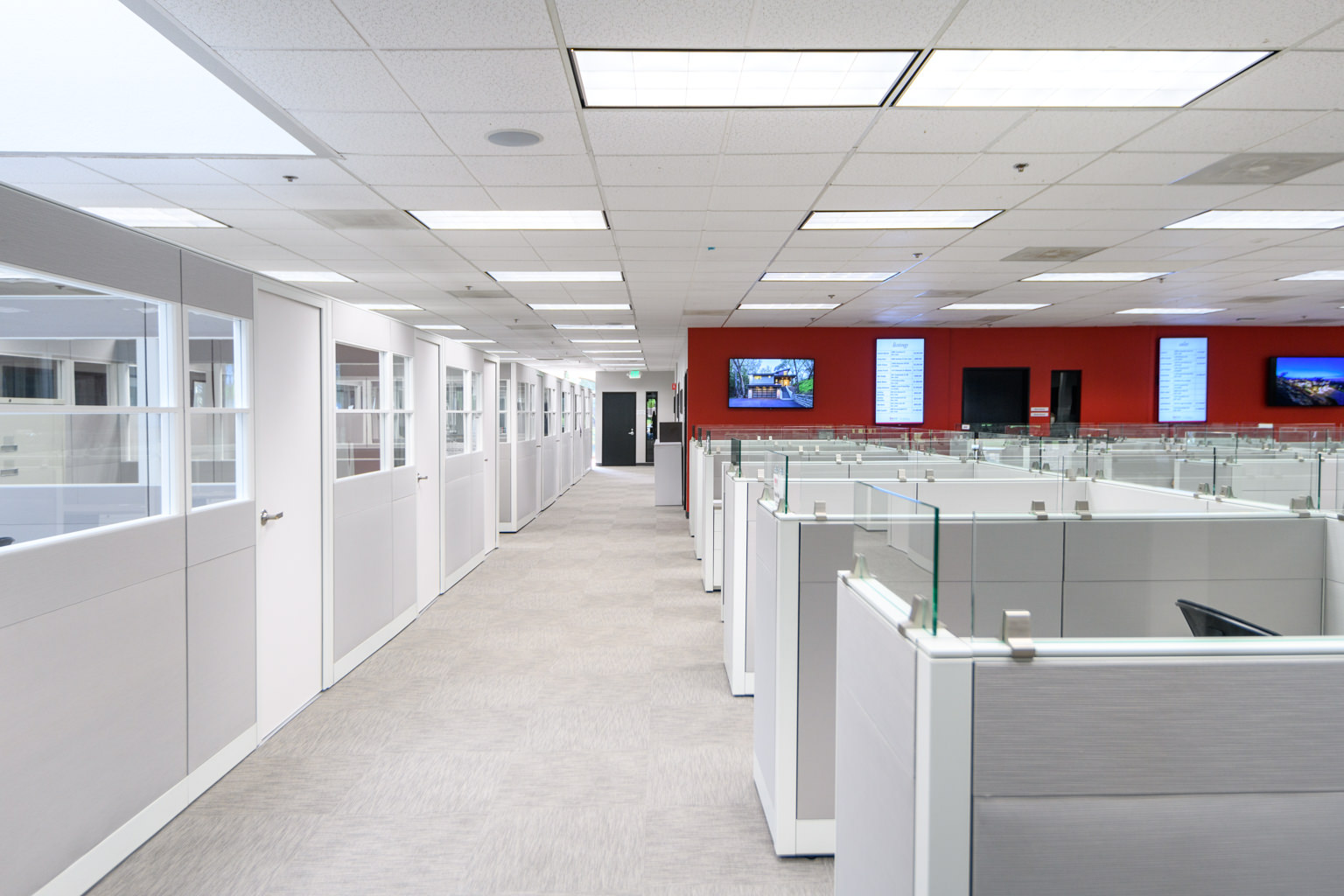 An open-plan office space featuring a long hallway lined with private office doors on the left and rows of modern cubicles on the right beneath a drop ceiling.